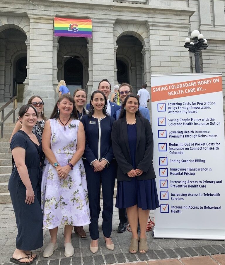 A group of CCHI staff, Colorado legislators, and additional advocates standing outside the Colorado Capitol with a post that lists ways they have saved Coloradan's money on health care.